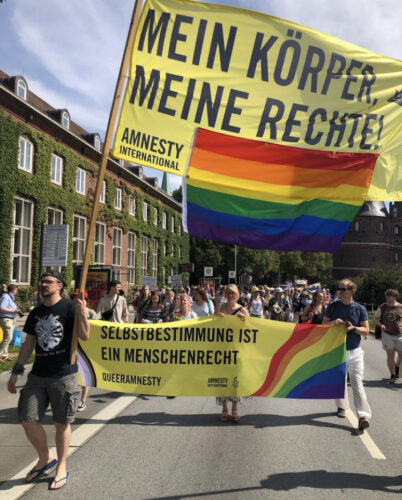 Zwei Amnesty Mitglieder tragen ein Banner, welches an Holzpfählen befestigt ist, um es hoch und sichtbar zu machen. Das Banner hat die Aufschrift „mein Körper meine Rechte“ und eine Regenbogenflagge unter dem Schriftzug befestigt. Hinter den zwei Amnesty Mitgliedern laufen weitere Amnestys mit dem Banner „Selbstbestimmung ist ein Menschenrecht“. Dies ist gelb und hat recht einen wellenartig seitlich verlaufenden Regenbogen und links ebenfalls wellenartige verlaufende Farben schwarz, braun, hellblau, hellrosa, weiß und wieder gelb mit einem lilafarbenen Kreis. Dahinter sind sehr viele weitere CSD Teilnehmer*innen.