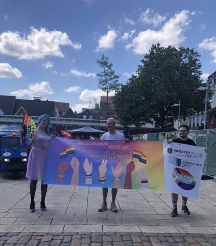 Drei Personen halten ein Banner mit der Aufschrift „zusammen vereint Christopher Street Day Stade“. Unter dem Schriftzug sind Hände verschiedener Hautfarben zu sehen mit Pride Flaggen Bändchen um. Der Hintergrund des Banners ist in Regenbogenfarben gehalten. Links hält das Banner eine Drag Queen im rosa Kleid mit einer türkis-blauen Perücke, welche sie zu einem geflochtenen Zopf gebunden hat. Sie hält in ihrer rechten Hand einen großen bunten Fächer. Hinter den Personen sind Gebäude zu sehen und ein blauer Himmel.