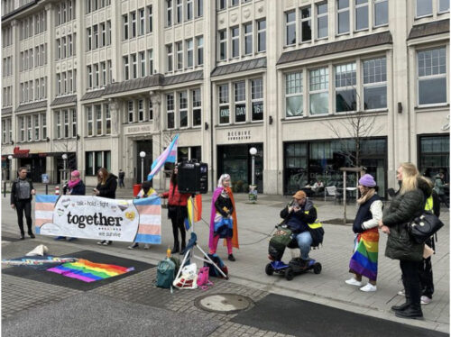 Hier sind Queeramnesty Mitglieder und enbygalactic:transtastic Mitglieder bei einer Kundgebung zum Trans Remembrance Day zu sehen. Vier Personen halten ein Banner mit der Aufschrift „take hold fight together we survive“. Es hat eine Transflagge rechts und links abgebildet und über dem Schriftzug kleine Herzen aus anderen Flaggen (agender, genderqueer, trans, nichtbinär und intersex). Vor dem Banner liegt eine Regenbogenflagge und eine Disability Pride Flagge. Rechts vom Banner weht eine weitere Transflagge. Eine Rednerin trägt eine Lesbenflagge und eine AroAce Flagge um den Körper. Ein weiterer Redner sitzt in einem E-Scooter daneben mit einem Mikrofon und einem Smartphone zum Ablesen in der Hand. Neben ihm steht eine weitere Rednerin mit einer Regenbogenflagge um die Hüfte. Sowie zwei weitere Teilnehmer*innen. 