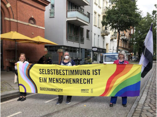 Drei Amnesty Mitglieder halten das Banner „Selbstbestimmung ist ein Menschenrecht“. Das Mitglied rechts außen hält zusätzlich eine Stange mit der Asexuellen Flagge. Das Foto wurde aufgenommen in der Straße Lange Reihe.