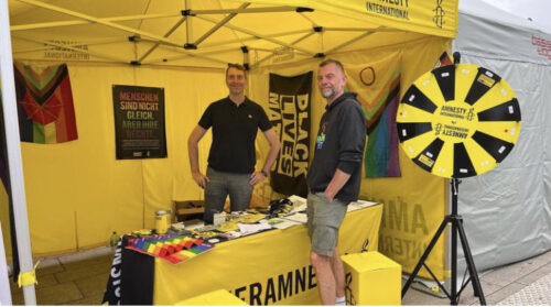 Zwei Amnesty Mitglieder stehen um einen Tisch mit Merchandise von Amnesty unter einem gelben Zelt. Der Tisch hat ein gelbes Banner an der Vorderseite mit der Aufschrift „Queeramnesty“ in schwarz. Neben den Mitgliedern ist ein Glücksrad in schwarz und gelb mit Nummern drauf. Im Zelt hängen mehrere Flaggen (Black Lives Matter Flagge, Progress Pride Flagge mit Inter- und Progress Pride Flagge mit einem roten Regenschirm für die Sexarbeiter*innen).