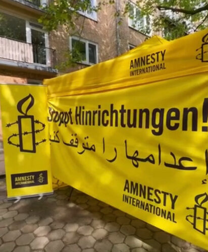 Amnesty Infostand mit einem Banner auf dem "Stoppt Hinrichtungen" auf deutsch und arabisch geschrieben steht. Daneben ein Aufsteller mit dem Amnesty-Logo.